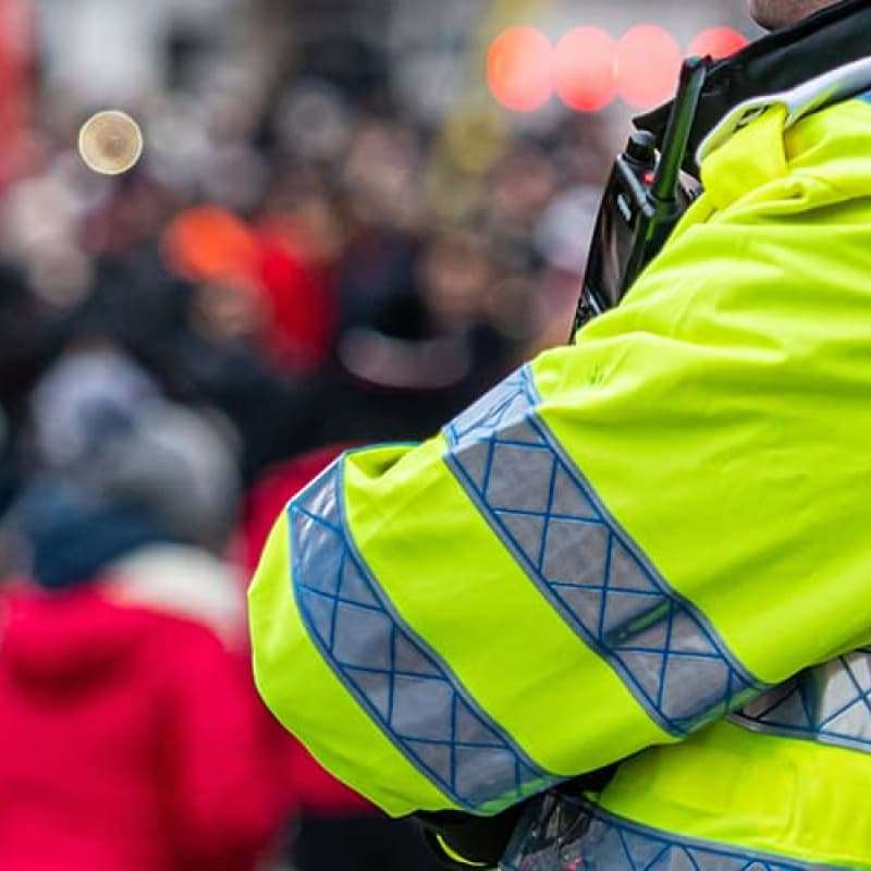 Police man in fluorescent jacket standing with his arms cross in-front of a crown that is blurred out | e-Surgery