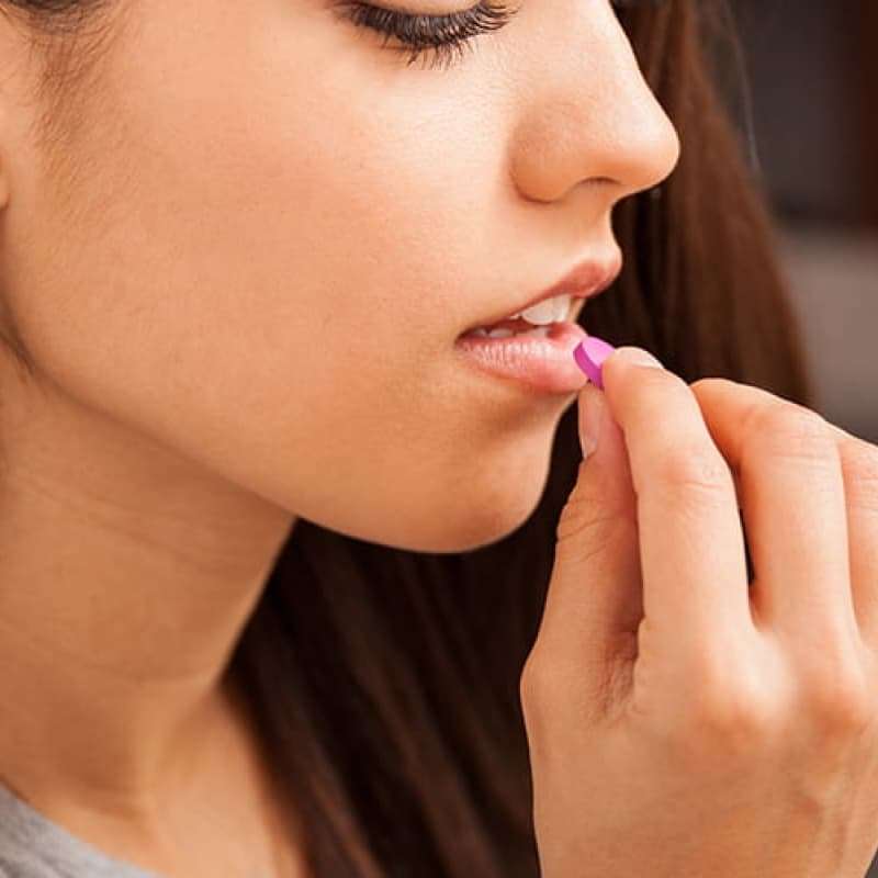 Myth busting emergency contraception, young woman with long brown hair sitting on a sofa placing a pill into her mouth. She has a glass of water in her other hand | e-Surgery