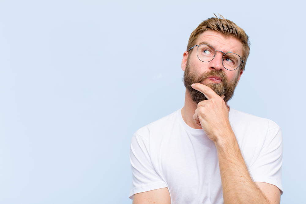 Young male with a bears wearing glasses and a white t-shirt looking interested. He has his chin rested in his thumb and index finger | The Male Pill: Myth Or The Future Of Contraception? | e-Surgery