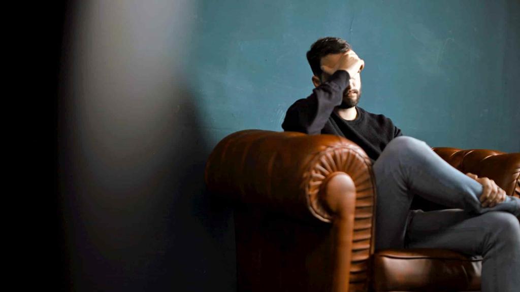 Middle aged man sitting on a brown leather sofa resting his head into his left hand with his elbow on the arm rest | Tired All The Time? 6 Common Anaemia Symptoms | e-Surgery