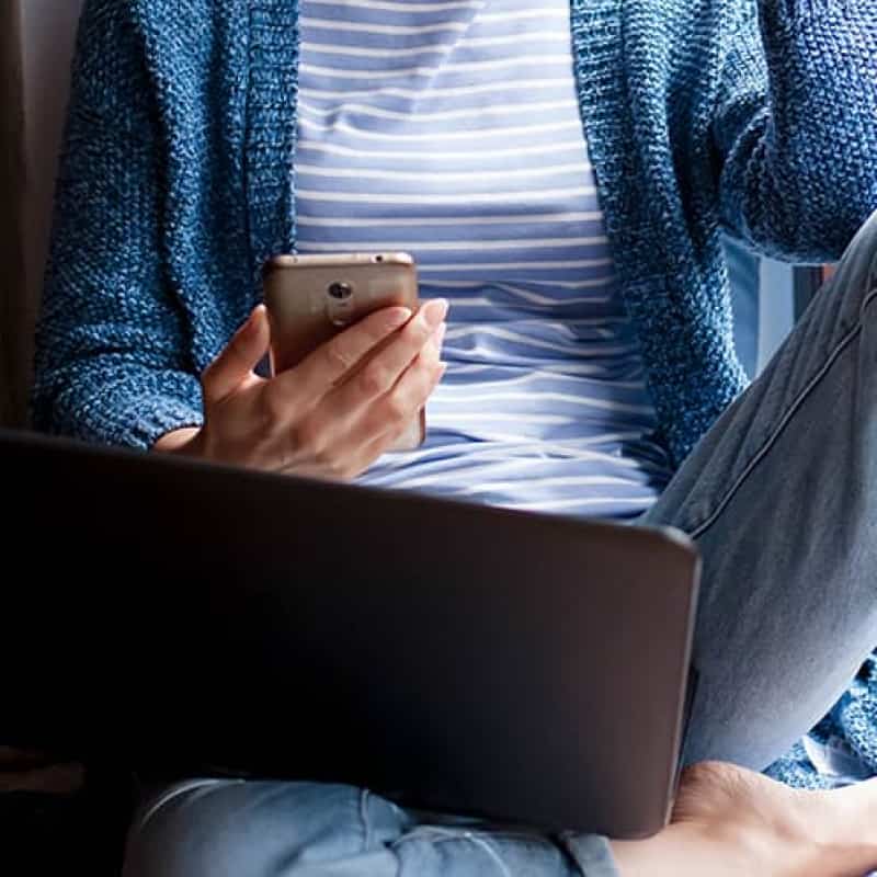 Woman sitting by window holding phone and laptop, ways to benefit your well being in lockdown, e-Surgery