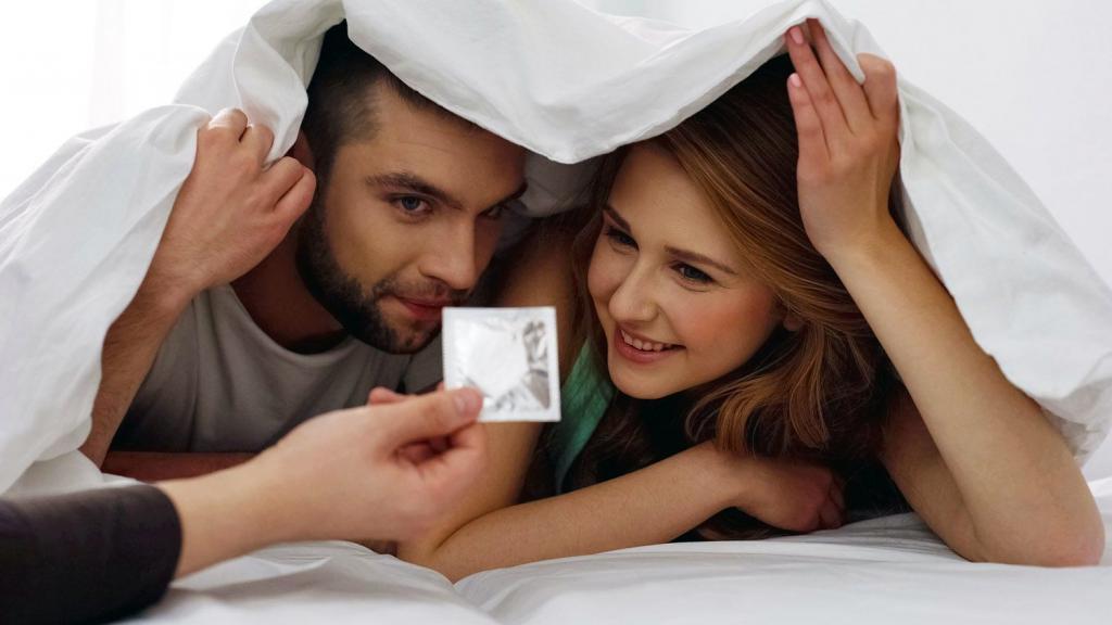 Young couple hiding under the covers. Both are peeking out the bottom of the duvet looking at a hand that is passing them a condom | Why Do Young People Keep Their STIs a Secret? | e-Surgery