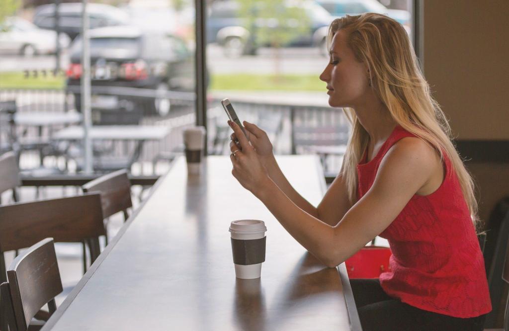 Young blonde girl in a coffee shop sitting at a table on her phone. It is suggested that she is using a period-tech application to track her period | Period-tech: 5 Ways Technology is Changing Women’s Health | e-Surgery