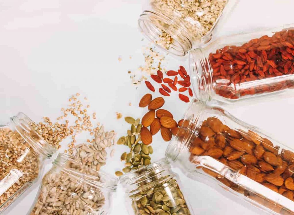 Jars of seeds and nuts laying on their side without lids. The seeds and nuts are spilling out onto the white table cloth. The image has been taken from an aerial view | Does Your Diet Trigger Migraines? | e-Surgery