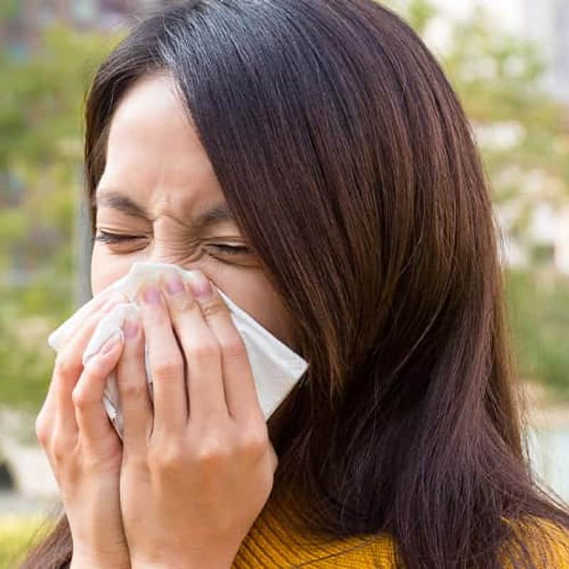 Finding the best nasal spray for hay fever, woman in yellow jumper sneezing into a tissue | e-Surgery